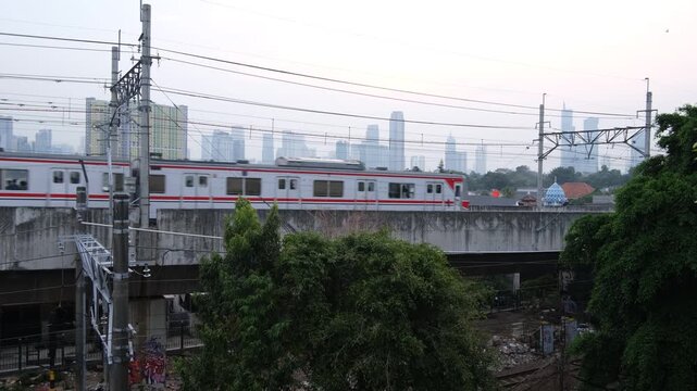 A commuter train travels on an elevated railway track with power lines above. The scene is set in an urban environment with high-rise buildings faintly visible in the background under a cloudy sky.  

