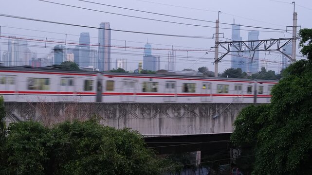 A commuter train travels on an elevated railway track with power lines above. The scene is set in an urban environment with high-rise buildings faintly visible in the background under a cloudy sky.  
