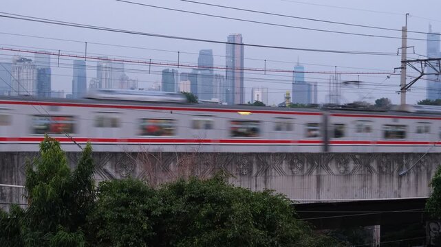 A commuter train travels on an elevated railway track with power lines above. The scene is set in an urban environment with high-rise buildings faintly visible in the background under a cloudy sky.  
