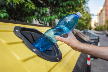 Hand recycling a blue plastic bottle into a yellow container on a city street in Rome, Italy, promoting waste sorting and environmental care.
