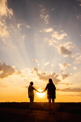 Fototapeta premium Two women holding hands in silhouette at sunset with dramatic rays of light breaking through the clouds