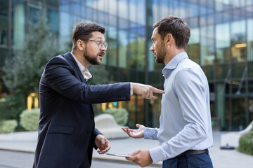 Two angry businessmen are having a tense argument, one pointing a finger while the other gestures in disagreement, depicting workplace stress and conflict outside a modern office building