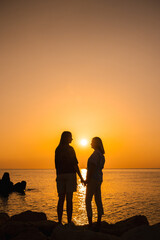 Fototapeta premium Two women standing on rocks by the sea at sunrise, holding hands and facing each other, symbolizing love, family, emotional wellbeing, and a deep bond with nature