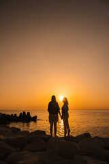 Fototapeta premium Two women standing hand in hand on coastal rocks at sunrise, gazing at the sea horizon, symbolizing family bonds, emotional health, love, and peaceful connection with nature