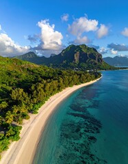 Aerial view of a pristine, crescent-shaped beach curving along turquoise water, backed by lush greenery and dramatic mountains under a vibrant blue sky
