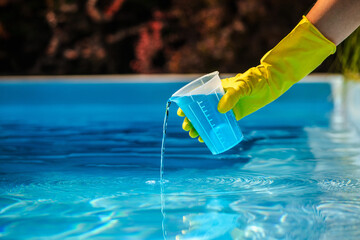 Hand with yellow protective glove pouring blue chemical liquid from measuring cup into swimming pool water for cleaning and disinfection