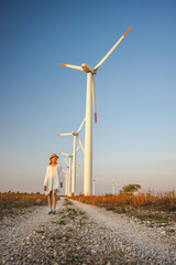 Obraz premium Young woman in white dress and hat walking along a gravel path near wind turbines at sunset.
