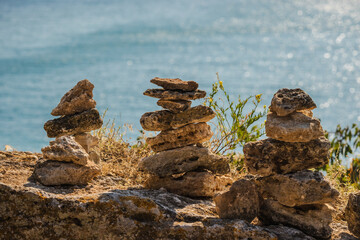 Three stacks of balanced stones on a rocky cliff overlooking the blue sea, dry grass and small plants growing nearby.