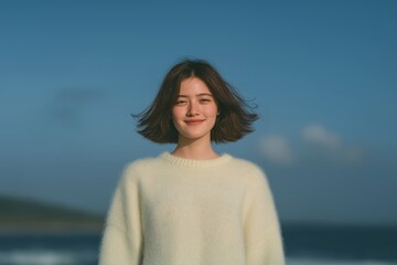 Obraz premium Natural portrait of a cheerful young woman with a short bob and a cream sweater, smiling with a deep blue sky and blurred coastline in the background