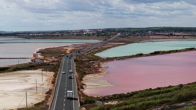 Aerial view of road crossing colorful salt lagoons near Santa Pola, Alicante, Spain. Traffic, old house, and pink and turquoise ponds under cloudy sky.