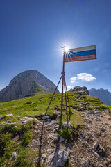 Scenic view from the  sun backlit Slovene flag on Kamniško sedlo in the Kamnik-Savinja Alps, Slovenia, showcasing alpine peaks like Brana and Turska gora, summer landscape