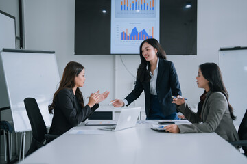 Three young Asian businesswomen collaborating and analyzing financial charts and graphs on a display screen in a modern conference room, demonstrating teamwork and professional discussion