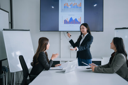 Asian businesswoman presenting sales performance data and charts on a large screen during a corporate meeting in a modern office, engaging with her female colleagues