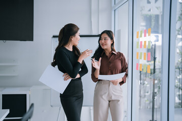 Two Asian businesswomen stand by a glass wall covered in colorful sticky notes, collaborating on creative brainstorming and project planning with focused, positive engagement