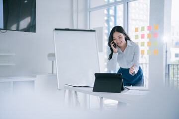Young Asian businesswoman on a phone call, smiling and giving a thumbs up while reviewing a tablet in a bright, modern office — confident, professional, productive
