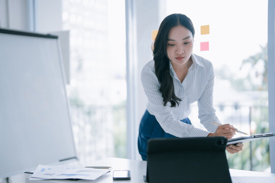 Asian businesswoman standing at a desk, reviewing a clipboard with documents, and using a digital tablet, demonstrating focus on business analysis and professional planning