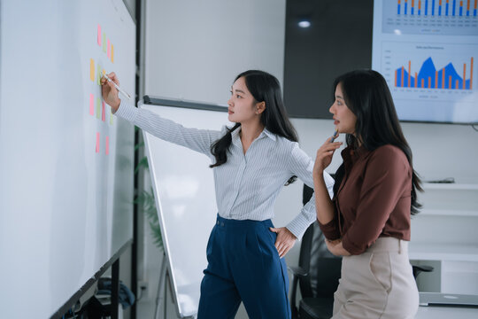 Two young businesswomen are brainstorming ideas and analyzing data, collaborating on a whiteboard with sticky notes while developing innovative strategies during an office meeting - Powered by Adobe