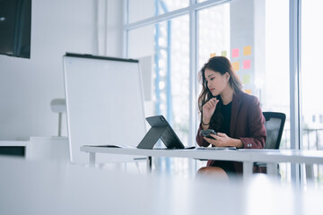 Young Asian businesswoman concentrating on tasks, analyzing data on her tablet, and using her smartphone for communication in a modern, bright office environment