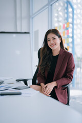 Asian businesswoman in smart professional attire smiles confidently at her desk with documents and phone, projecting leadership, success and a positive modern office presence