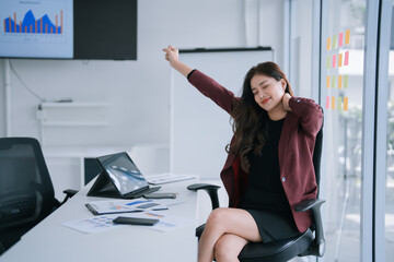 Asian businesswoman stretching neck and shoulders while sitting at her office desk, taking a well-deserved break from work, indicating stress relief and a need for relaxation