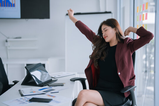 Asian businesswoman, comfortably seated at her modern office desk, feeling satisfied while stretching her arms and taking a refreshing break from work, indicating relaxation and self-care