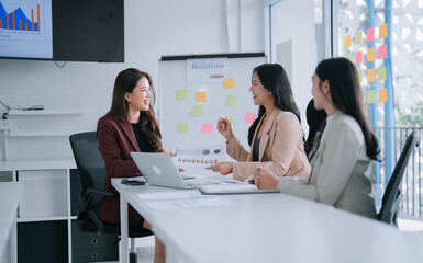 Three young Asian businesswomen are collaborating, analyzing financial charts and data on documents and a laptop during a corporate meeting in a modern office, focusing on teamwork and strategy
