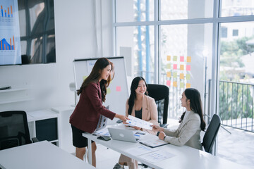 Three Asian businesswomen are collaborating, analyzing, and discussing a marketing report during a corporate meeting in a modern office, one passing documents to her colleague while smiling