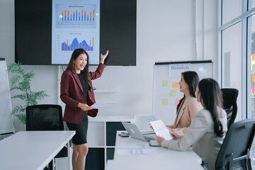 Businesswoman presenting sales performance charts on a large screen during a corporate meeting, actively engaging with two female colleagues who are attentively observing the data