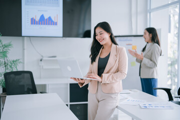 Asian businesswoman smiling while holding a laptop, reviewing data and charts displayed on a conference screen during a professional business meeting with a colleague presenting in the background