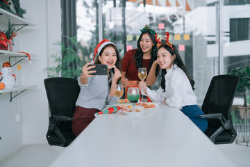 Three smiling young Asian women celebrating an office Christmas party, wearing Santa hats and reindeer antlers, taking a selfie with a smartphone while enjoying food and drinks