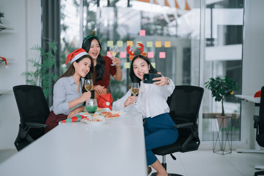 Three young Asian businesswomen are raising glasses of wine, wearing Christmas hats and headbands, and taking a selfie at a festive office party, celebrating holidays together