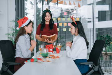 Three Asian female colleagues at a festive office Christmas party, wearing Santa hats and...