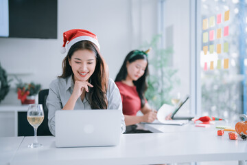 Young Asian businesswomen in Santa hats and festive headbands smile while working on laptops in a modern office, blending holiday cheer with professional teamwork and productivity