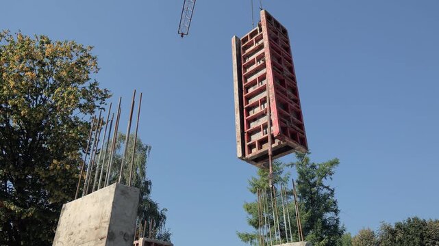 Large red formwork panel lifted by crane at construction site, concrete columns with steel rebars visible against clear blue sky and surrounding trees
