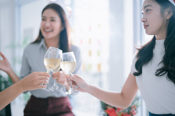 Asian women friends raising wine glasses in a cheerful indoor holiday party, smiling and toasting together in a festive office or social gathering celebrating success and friendship