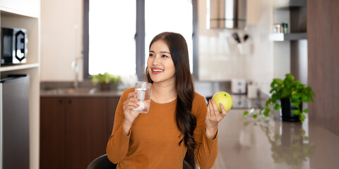 Asian attractive female in kitchen holding green apple and a glass of water drinking fresh water after wake up in morning for health.