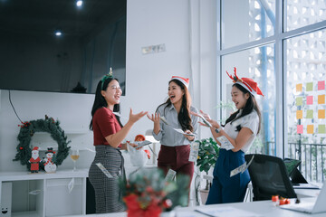 Three happy Asian businesswomen, wearing festive hats, celebrating an office Christmas party,...