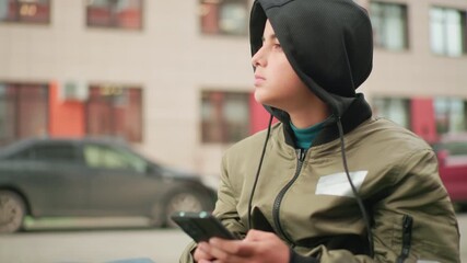 Teen boy in hoodie jacket holding smartphone looks to background with parked cars and residential building behind, appearing curious and observant while seated outdoors in urban setting - Powered by Adobe