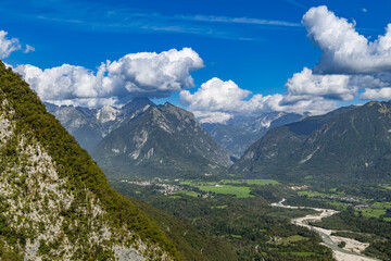 A view of Soča Valley near Žaga village, Bovec, Slovenia, with an emerald river, lush greenery, and majestic Alps under a vibrant blue sky with dramatic clouds