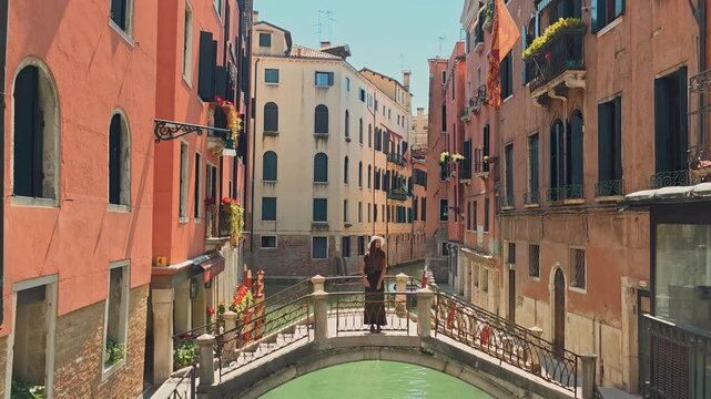 Woman posing on Venetian bridge over canal. A stylish woman in a hat stands on a small arched bridge in Venice, surrounded by colorful buildings and a peaceful canal.