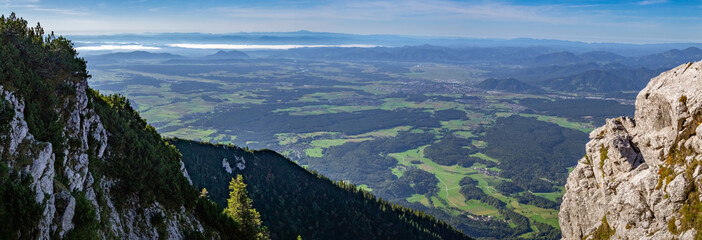 Panoramic view from Storžič mountain towards Ljubljana, Slovenia, with lush green fields, forested slopes, and rocky cliffs under a hazy blue sky.