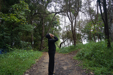 A solitary figure, dressed in dark attire, stands on a dirt path deep within a lush, temperate forest.