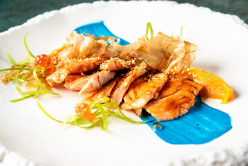 Gourmet teriyaki chicken with sesame seeds, bonito flakes, salmon roe, and citrus garnish on vibrant blue sauce. Modern fusion cuisine plated on white ornate dish