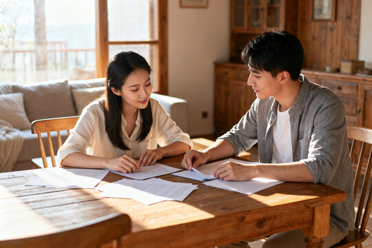 A young couple reviews documents together at a wooden table in a cozy, sunlit home. - Powered by Adobe