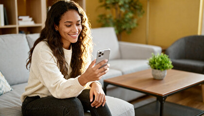 Woman with phone on sofa. Happy beautiful multiethnic girl using smartphone at home. Young person relaxing in cozy living room, browsing social media, texting. Lifestyle technology.