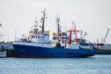 A large blue and white trawler-style fishing boat is moored at a quay in a harbor against a bright, overcast sky.