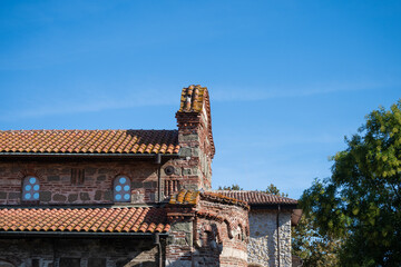 A historic Orthodox church structure in Nesebar, characterized by a mix of red brick, stone masonry, and terracotta tile roofs, is bathed in sunlight.