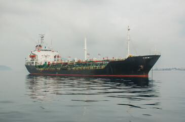 A large tanker vessel rests on the still sea in Ternate, Indonesia, with a volcanic island in the background. A symbol of global trade, energy transport, and the maritime industry