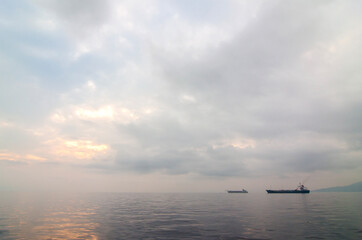 Fototapeta premium A large cargo ship is anchored on the exceptionally calm waters of Ternate, Indonesia, at dawn or dusk. A peaceful and minimalist scene with the sea reflecting the soft, cloudy sky.