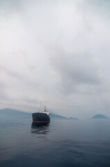 A large cargo ship sails in the waters of Ternate, Indonesia, with the majestic Mount Gamalama volcano in the background. A dramatic scene of an active volcano towering over the sea